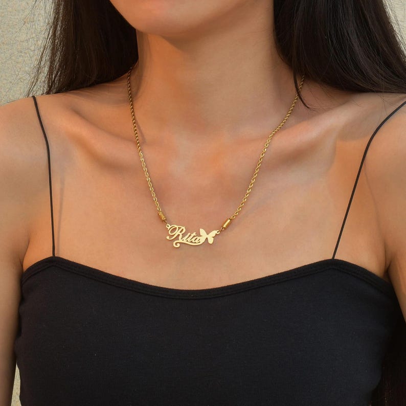 Pu&ograve; includere: Una collana color oro con catena a corda e un ciondolo personalizzato con il nome Rita scritto in corsivo, accompagnato da un piccolo dettaglio a forma di farfalla ritagliata fissato sul lato destro della targhetta.