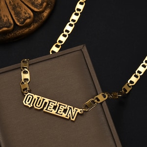 Pu&ograve; includere: Una collana color oro con un ciondolo centrale a targhetta con la parola QUEEN in un carattere in grassetto in stile blocco. Il ciondolo &egrave; fissato a una catena a maglie larghe e piatte che si estende verso l'alto, mostrando una finitura metallica lucida.