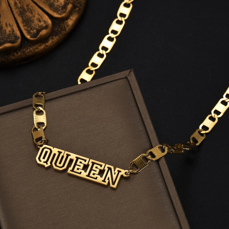 Pu&ograve; includere: Una collana color oro con un ciondolo centrale a targhetta con la parola QUEEN in un carattere in grassetto in stile blocco. Il ciondolo &egrave; fissato a una catena a maglie larghe e piatte che si estende verso l'alto, mostrando una finitura metallica lucida.