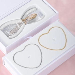 May include: A white box containing nail clippers and two heart-shaped necklaces. The clippers have gold blades and white handles. One necklace is gold, the other is silver.