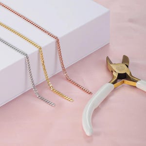May include: Three metal chain necklaces in silver, gold, and rose gold are displayed on a white surface. A gold and white wire cutter tool is positioned on a pink background. The chains are likely for jewelry making.