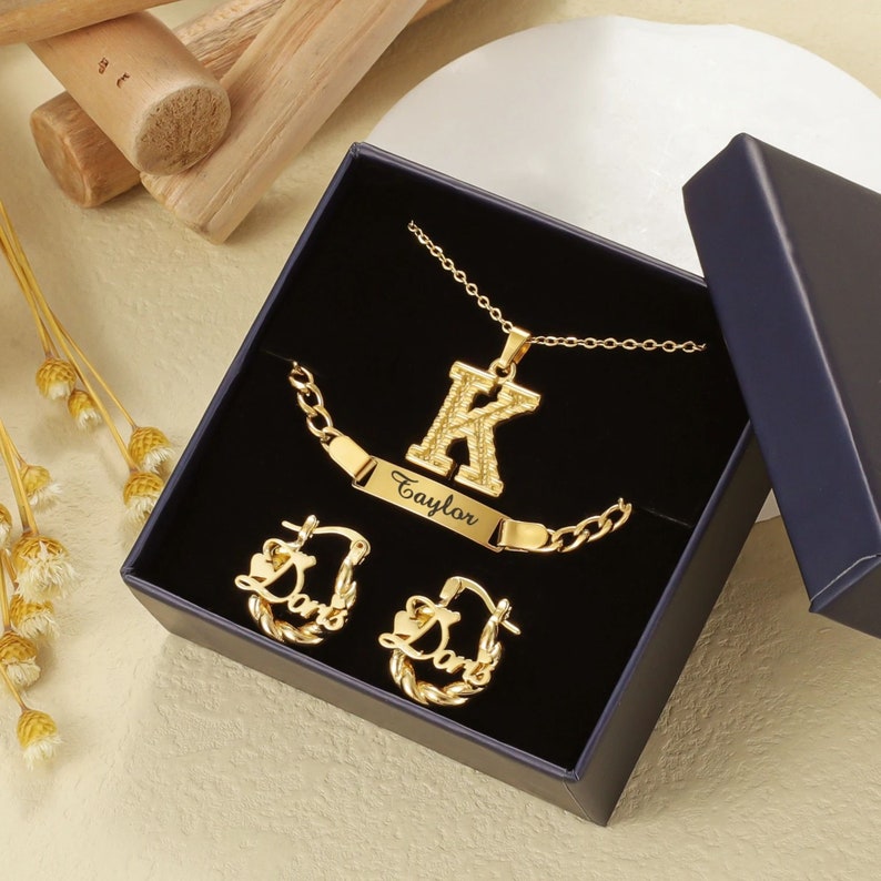 May include: Gold jewelry set in a black box. The set includes a necklace with a letter K pendant, a bracelet with a nameplate that says "Taylor", and a pair of hoop earrings with the word "Love" on them.
