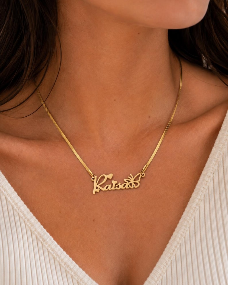Pu&ograve; includere: Una collana color oro con una catena piatta a spina di pesce e un ciondolo con nome personalizzato che riporta la scritta Raisa in corsivo. Il ciondolo include un piccolo cuore sopra la lettera i e una sagoma di farfalla sul lato destro.