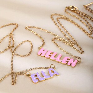May include: Gold-colored chain necklaces with name pendants. One pendant spells "HELLEN" in pink letters with a gold outline. Another pendant spells "ELLA" in purple letters with a gold outline. The necklaces are displayed on a light-colored surface.