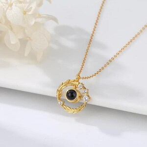 Puede incluir: Un collar de oro con un colgante redondo que presenta una piedra negra rodeada de peque&ntilde;as piedras transparentes. El colgante tiene forma de corona de laurel con hojas y tiene una cadena delicada.