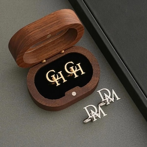 Op de afbeelding: Een houten sieradendoosje met goudkleurige manchetknopen binnenin, met de letters "GH". Naast de doos liggen zilveren manchetknopen met de letters "DM". De doos is open en onthult een zwart fluwelen interieur.