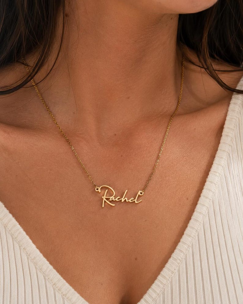 May include: A gold-toned metal necklace featuring a delicate chain and a central pendant displaying the name Rachel in a cursive script font. The nameplate is integrated directly into the chain, creating a continuous, lightweight piece of jewelry.