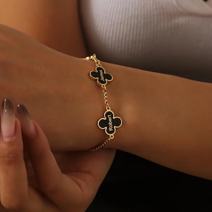Könnte beinhalten: Ein goldenes Kettenarmband mit schwarzen Kleeblatt-Anhängern. Jeder Anhänger trägt einen eingravierten Namen: "James" und "Robert".
