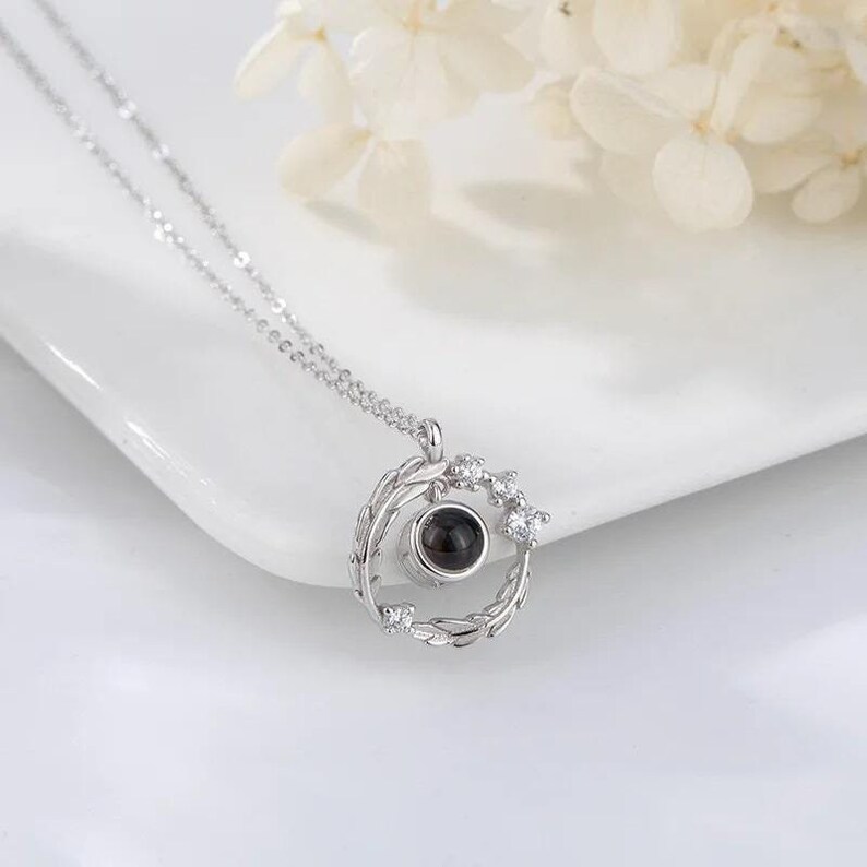 Puede incluir: Collar de plata con un colgante circular que presenta una peque&ntilde;a piedra negra rodeada de una corona de hojas y peque&ntilde;as piedras transparentes. El colgante proyecta un mensaje cuando se ve a trav&eacute;s de la piedra.