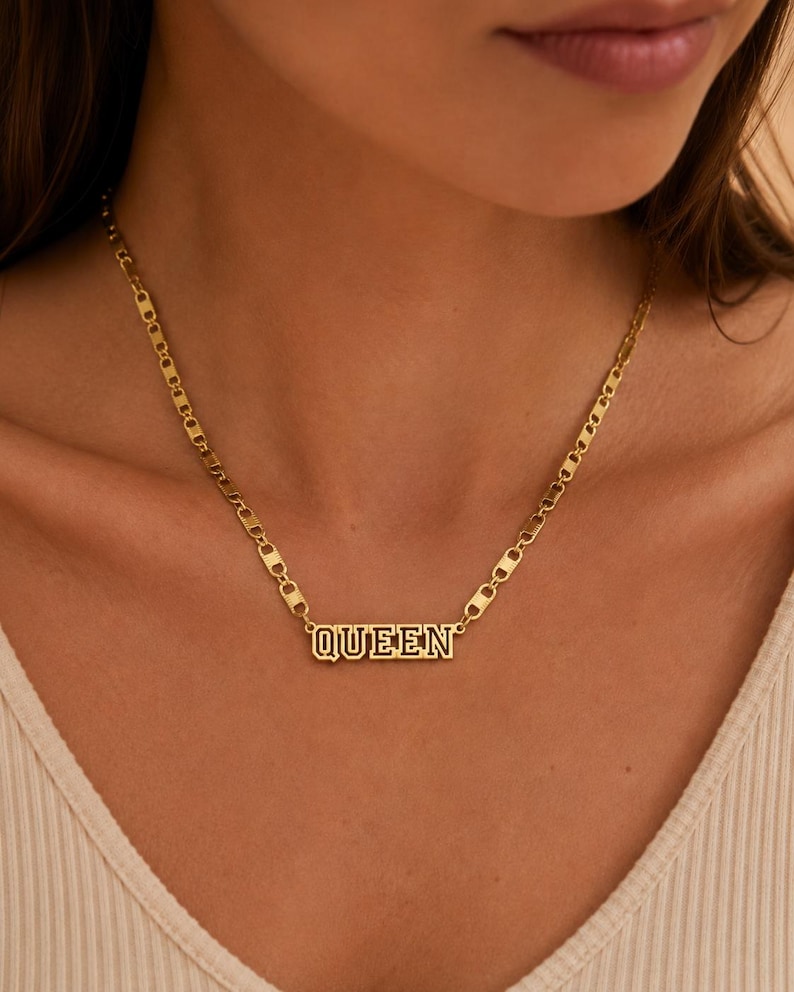 Pu&ograve; includere: Una collana color oro con un ciondolo rettangolare che riporta la parola QUEEN in un carattere a blocchi. Il ciondolo &egrave; fissato a una catena composta da maglie piatte, allungate e arrotondate che si uniscono per formare un design metallico strutturato.