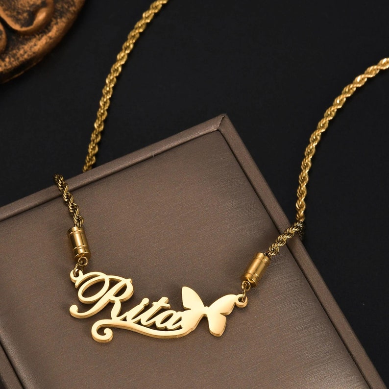 Pu&ograve; includere: Una collana color oro con catena a corda e un ciondolo con il nome Rita scritto in corsivo, accompagnato da una piccola sagoma di farfalla fissata sul lato destro della scritta.