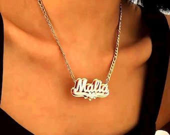 Può includere: Una collana con targhetta color oro che riporta il nome Malia in carattere corsivo. Il ciondolo presenta un design a strati con una superficie testurizzata scintillante e un piccolo dettaglio a forma di cuore, appeso a una catena figaro dorata.