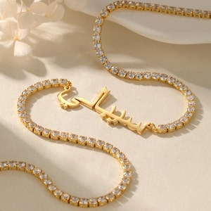 May include: Gold-toned necklace with a name pendant in Arabic script. The necklace features a tennis chain design with clear, square-cut stones. The pendant is in a stylized font. The jewelry is displayed on a neutral background.