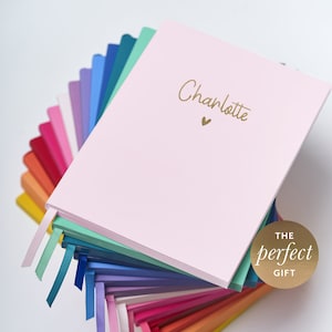 May include: A pink notebook with the name "Charlotte" written in gold lettering and a small gold heart. The notebook is part of a stack of notebooks in various colours with ribbon bookmarks.