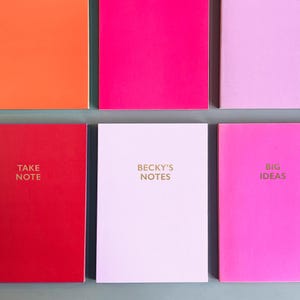 May include: Six colourful notebooks with different text on the front. The notebooks are arranged in a grid pattern. The top row has orange, pink, and light purple notebooks. The bottom row has red, light purple, and pink notebooks. The text on the notebooks reads "Take Note", "Becky's Notes", "Big Ideas".