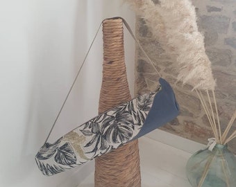 May include: A black and white yoga mat bag with a tropical leaf pattern and gold accents. The bag has a blue lining and a long strap. The bag is sitting on a wicker vase.