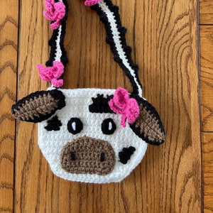 May include: A white crocheted cow-shaped purse with brown ears and a brown nose. The purse has black spots and pink flowers on the strap.