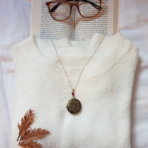 Peut inclure: Un pull blanc en fourrure avec un collier de chaîne en or et un pendentif rond orné avec une pierre rouge. Le collier est posé sur le pull. Il y a deux brindilles brunes sur le côté gauche du pull. Le pull est sur une surface blanche.