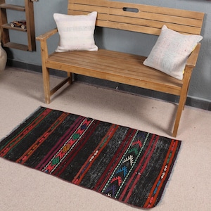 Peut inclure: Un tapis à motifs noirs et rouges avec des accents colorés. Le tapis est posé sur un sol beige clair devant un banc en bois avec des coussins.