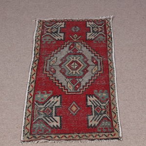 May include: A small, rectangular red rug with a central geometric design in gray, beige, and green. The rug has a patterned border and fringe on the short ends. The rug's dimensions are approximately 24 inches long and 12 inches wide.