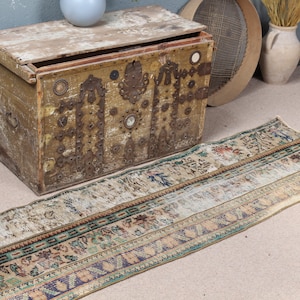 Puede incluir: Un cofre de madera antiguo con herrajes metálicos y un acabado envejecido se encuentra junto a una alfombra rectangular larga con un diseño descolorido. La alfombra presenta una mezcla de colores beige, azul y verde.