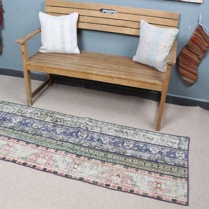 May include: A rectangular area rug featuring a multi-colored pattern with horizontal bands of blue, green, and muted red floral and geometric motifs. The rug is positioned on a light-colored floor in front of a wooden bench with two white pillows.
