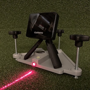 May include: A black Garmin device mounted on a tripod with three adjustable legs. The device is projecting a red laser line onto a green surface.
