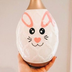 May include: White ceramic diffuser with a bunny face design. The diffuser has a gold top and a pink nose. The diffuser is labeled "Living".