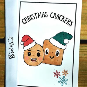 May include: A white greeting card featuring the text CHRISTMAS CRACKERS in black font. The illustration shows a round cracker and a square cracker, both with smiling faces and wearing festive Santa hats in red and green, with small snowflakes.