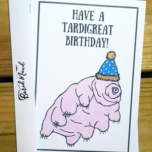 May include: A white greeting card featuring a pink tardigrade illustration wearing a blue polka-dot party hat with a yellow pom-pom. The text reads HAVE A TARDIGREAT BIRTHDAY! in black capital letters. The card is partially inside a white envelope.