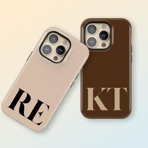 Peut inclure: Deux coques de téléphone, une beige et une marron, chacune avec des initiales différentes en noir et beige. La coque beige porte les initiales "RE", tandis que la marron affiche "KT". Les deux coques ont une finition brillante.