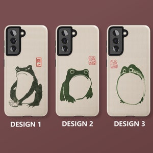 May include: Three phone cases with different designs featuring a frog illustration. The cases are beige with a black camera cutout and a black outline around the frog. The first case has a frog holding a bowl, the second case has a frog with its mouth open, and the third case has a frog with a circle around it.