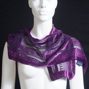 Può includere: Una sciarpa di velluto viola con un motivo geometrico bianco e nero. La sciarpa è realizzata in un tessuto morbido e lussuoso e ha un drappeggio delicato e fluido.