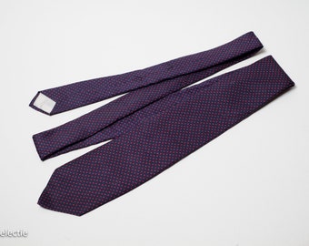 May include: A navy blue tie with a repeating pattern of small, red dots. The tie is laid out flat, showcasing its length and design. The fabric appears to be smooth and the overall look is classic and stylish.