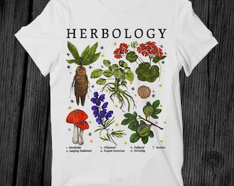 May include: White t-shirt with the word "HERBOLOGY" printed in black at the top. The shirt features a colorful illustration of various plants and mushrooms, including a mandrake, leaping toadstools, gillyweed, fanged geranium, puffapod, shrivelfig, and aconite.