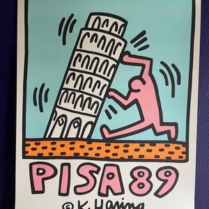 May include: A poster featuring a pink figure pushing the Leaning Tower of Pisa, with the text "PISA 89" in pink. The background is light blue, with orange and black accents. The artist's signature, K. Haring, is at the bottom.