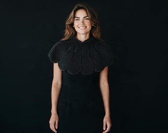 May include: A black collar accessory featuring a scalloped, petal-like design with intricate beadwork detailing. The collar is worn over a simple black garment, creating a textured, layered appearance with a high neckline and rounded edges.