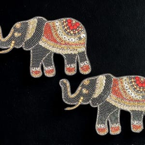 May include: Two beaded elephant patches. The elephants are black with gold, red, and white accents. The patches are embellished with beads and sequins. The elephants have their trunks raised and are facing left.