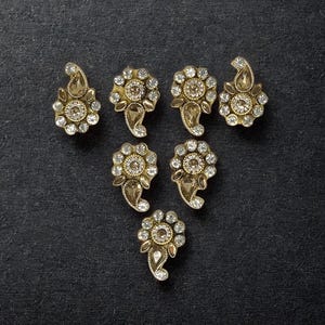May include: A set of seven decorative metal embellishments featuring a gold-tone finish, each shaped like a flower with a teardrop accent. The pieces are adorned with clear, faceted rhinestones and a central light brown stone in each floral centre.