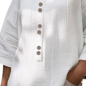 May include: A white long-sleeved shirt featuring a textured, crinkled fabric finish. The garment includes a vertical placket down the center front adorned with six round, metallic-toned buttons. The shirt has a simple round neckline and loose sleeves.