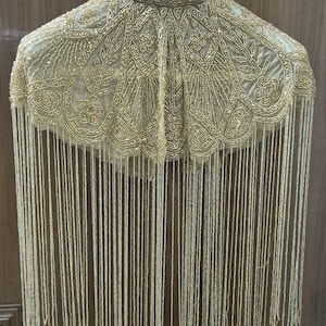 May include: A gold beaded capelet featuring a high collar and intricate floral patterns created with metallic beads. The garment is adorned with long, dangling gold beaded fringe that extends from the scalloped hemline, creating a layered effect.
