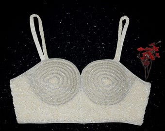 May include: A white, beaded bustier top with spiral detailing on the cups and thin, white straps. The bustier is embellished with small, shimmering beads and has a silver trim. A sprig of red flowers is to the right of the bustier against a black background.