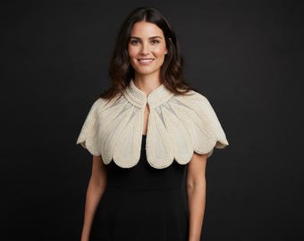 May include: A cream-colored, petal-shaped capelet featuring intricate rows of small, round, white beads along the scalloped edges and seams. The garment is worn over a simple black sleeveless dress against a dark background.