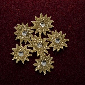 May include: A cluster of five star-shaped appliqués featuring gold-toned beadwork and a central clear circular rhinestone. Each star has twelve pointed rays, creating a textured, metallic appearance with a reflective centre on each individual piece.