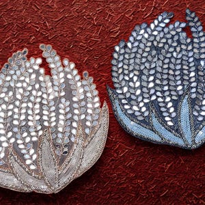 Può includere: Due oggetti decorativi a forma di fiore con intricati dettagli di specchi e paillettes. Uno è argentato e l'altro blu, entrambi su uno sfondo rosso strutturato. Il design assomiglia a un fiore stilizzato.