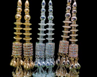 May include: Close-up of three decorative beaded tassels in gold, silver, and rose gold. Each tassel features a series of beads and a cylindrical section encrusted with rhinestones. The tassels are suspended from thin gold or silver cords.