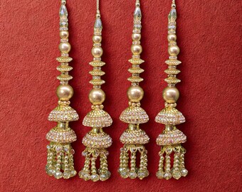 May include: Four gold-colored decorative tassels with intricate beadwork and crystal accents. Each tassel features a series of beads, a large central bead, and a beaded fringe. The tassels hang against a solid red background.