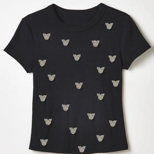 May include: A black short-sleeved t-shirt featuring a crew neckline and a repeating pattern of small, silver-toned, textured butterfly-shaped embellishments scattered across the front of the garment, creating a consistent decorative visual effect.