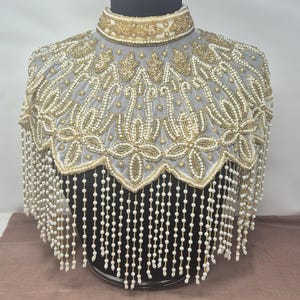 May include: A high-neck collar capelet featuring intricate gold beadwork and white pearl embellishments on a sheer fabric base. The garment displays a scalloped hemline adorned with long, dangling strands of white pearls that create a fringe effect.