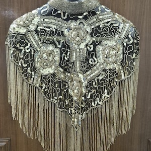 Peut inclure: Une capelette noire transparente ornée de perles dorées complexes et de motifs floraux à paillettes. Le vêtement possède un col haut et est terminé par de longues franges dorées pendantes sur le bord inférieur, formant une silhouette en V.